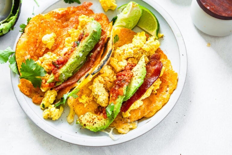 two breakfast tacos on a white plate, each taco is stuffed with eggs, crispy bacon, avocado slices and fresh salsa