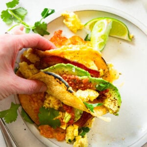 a hand holding a breakfast taco with eggs, bacon, cheese and avocado slices loaded in a corn tortilla