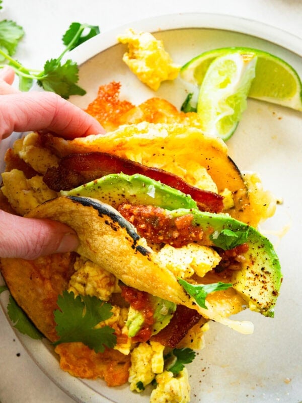 a hand holding a breakfast taco with eggs, bacon, cheese and avocado slices loaded in a corn tortilla