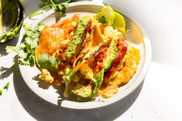 two breakfast tacos sitting on a white plate. each taco is loaded with eggs, bacon, avocado, cheese and salsa