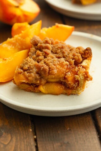 A slice of peach crumble bar on a white plate, topped with golden-brown streusel and served with fresh peach slices.