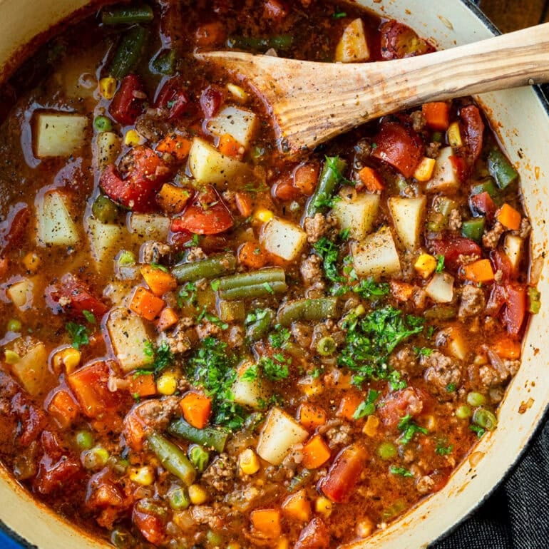 a large bot of ground beef vegetable soup topped with minced fresh parsley with a wooden spoon in the bowl