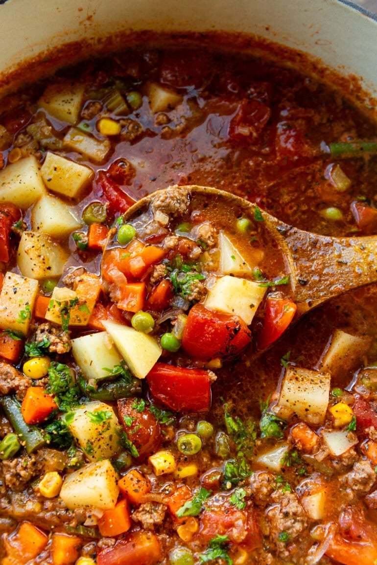 a wooden spoon listing a spoonful of ground beef and vegetables from a pot full of hamburger soup