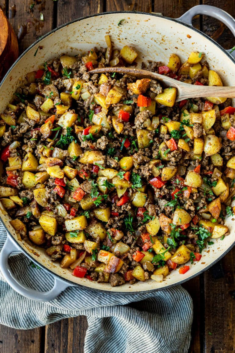 a photo of crispy potatoes, cooked ground beef, diced onions and red bell peppers topped with diced parsley in a cast iron skillet