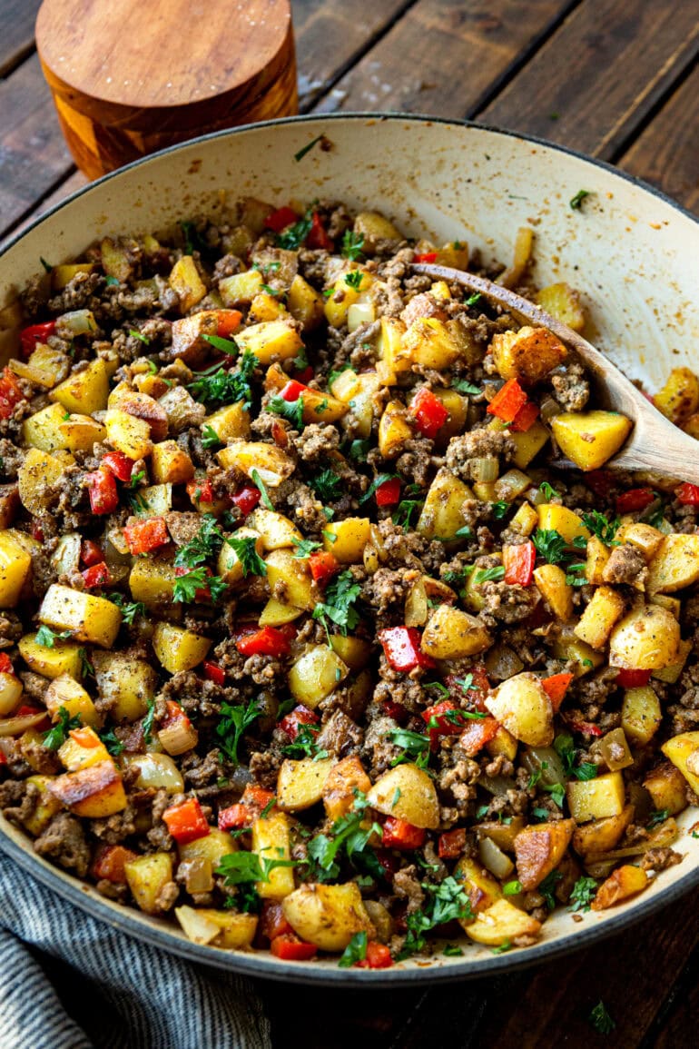 a photo of a cast iron skillet full of cooked ground beef and potatoes mixed with diced red bell peppers and onions and topped with mixed fresh parsley
