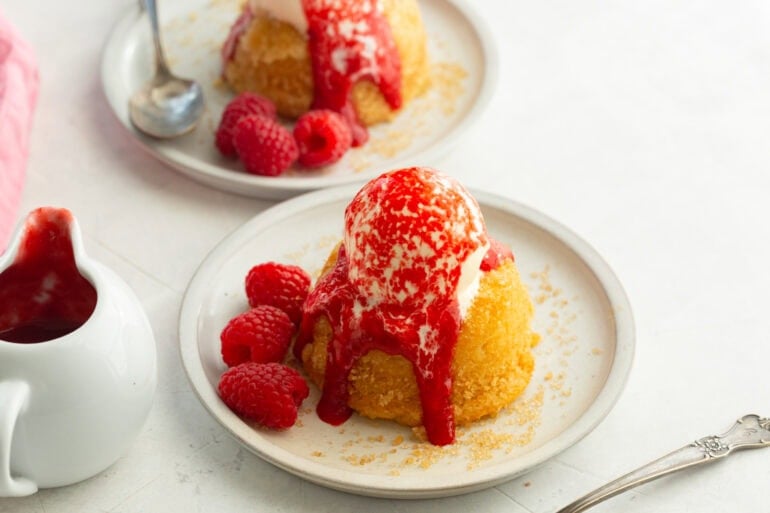 a gooey butter cake on a small dessert plate topped with melting vanilla ice cream and fresh raspberry sauce