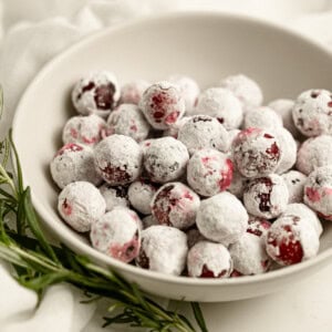 a small bowl of sugar coated whole cranberries with a sprig of fresh rosemary laying next to it.