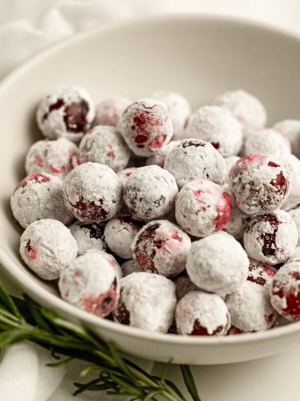 a small bowl of sugar coated whole cranberries with a sprig of fresh rosemary laying next to it.