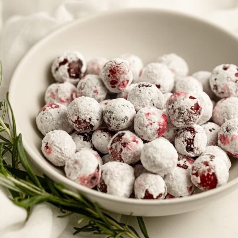 a small bowl of sugar coated whole cranberries with a sprig of fresh rosemary laying next to it.