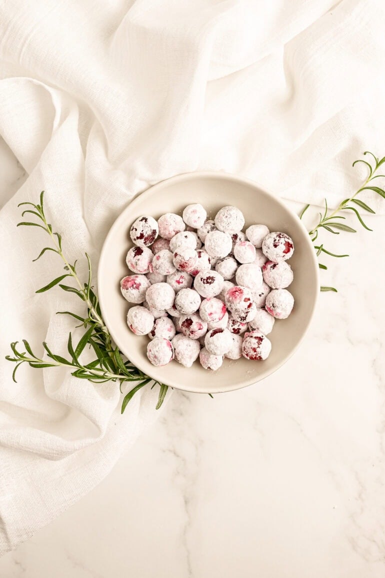 a small bowl full of candied whole cranberries with sprigs of fresh rosemary sticking out from under the bowl all on a white tablecloth