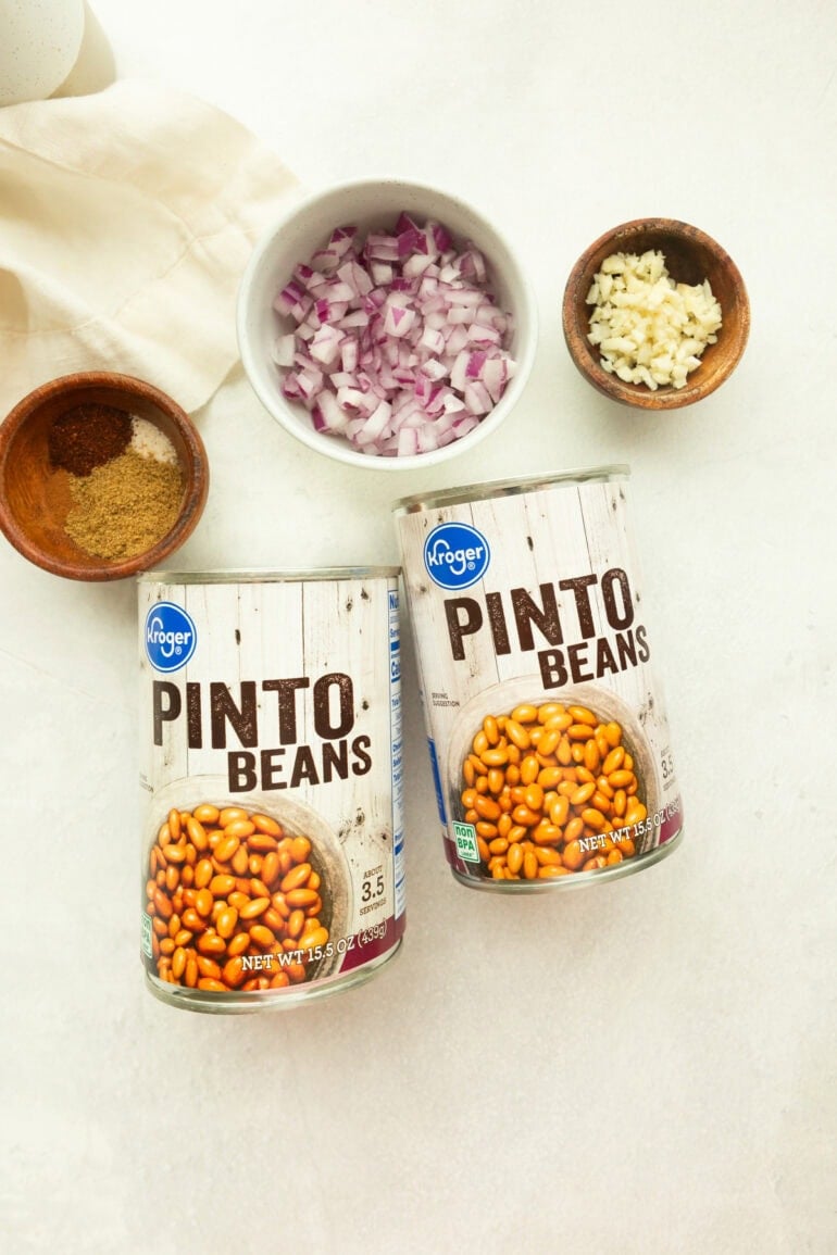 all the ingredients for canned pinto beans including two cans of pinto beans with minced red onions and garlic and several seasonings in a small bowl