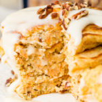 A photo of a stack of four carrot cake pancakes with a thick white cream cheese sauce poured over the top with a few chopped pecans and shredded carrots on top.