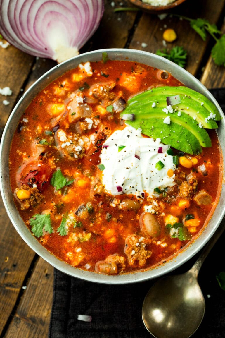 a large bowl of charro bean soup with pinto beans, corn, bacon, chorizo and topped with sour cream, sliced avocado, cilantro and diced red onions