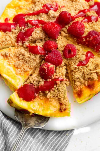 a photo of a baked yogurt sitting on a serving tray with a slice being removed topped with fresh strawberries and a graham cracker crust crumble