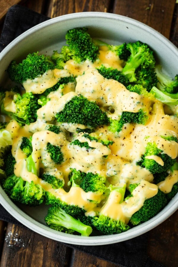 a large bowl of bright green steamed broccoli topped with cheddar cheese sauce and salt and pepper
