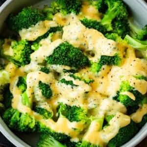 a photo of a large bowl of bright green steamed broccoli topped with a creamy cheese sauce and sprinkled salt and pepper
