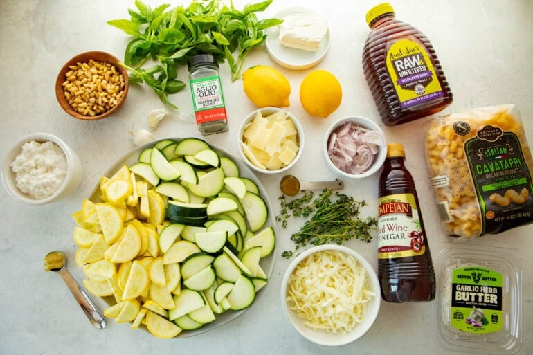 all the ingredients for cheesy zucchini pasta including shallots, pine nuts, fresh basil and thyme, zucchini and yellow squash and cavatappi pasta