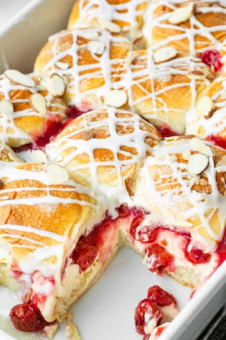 a photo of a baking dish full of golden baked sliders full of cherry pie filling and creamy cheesecake filling and topped with a drizzle and sliced almonds