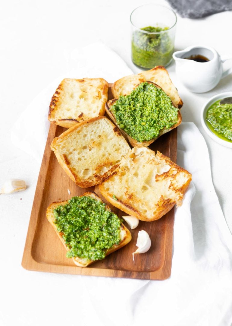 a photo of 3 ciabatta rolls sliced in half and toasted sitting face up on a wooden serving tray and 2 of the 6 slices have pesto spread on them.