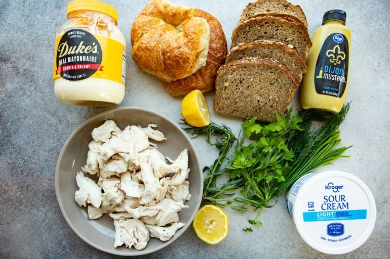 Ingredients like a jar of mayonnaise, dijon mustard, sour cream, lemon, fresh herbs, rotisserie chicken, croissants, and yummy seedy bread. 