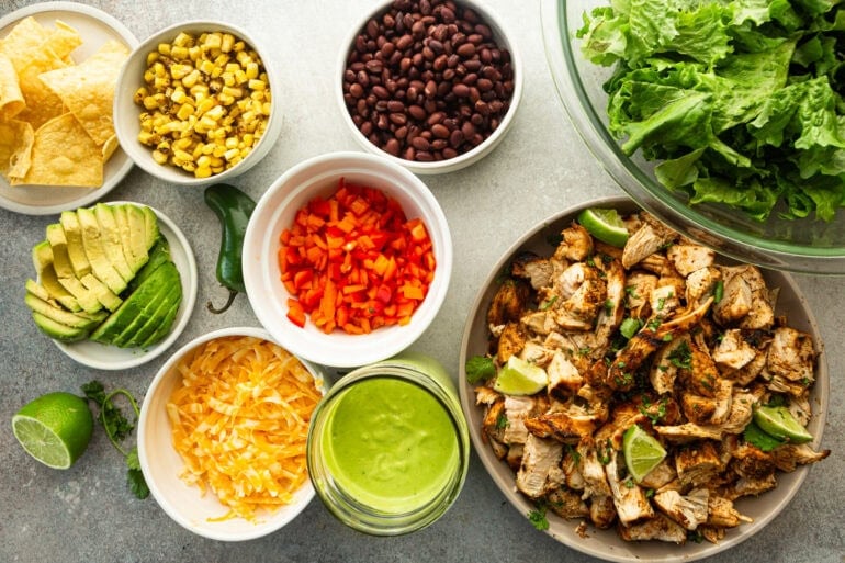all the ingredients for chicken taco salad in separate smaller bowls including grilled chicken chunks, cheese, lettuce, tomatoes, black beans, dressing, corn and avocado