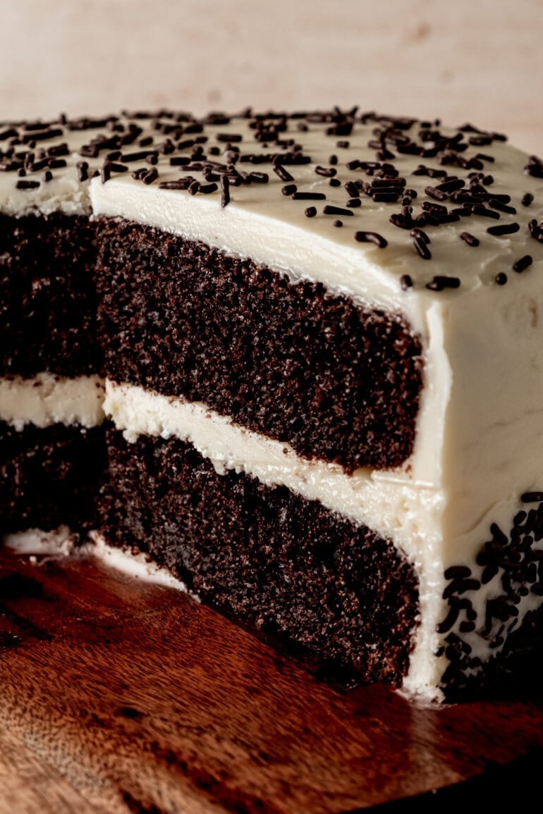 a two layer chocolate cake with vanilla buttercream frosting topped with chocolate sprinkles and slice cut out of it so you can see the inside