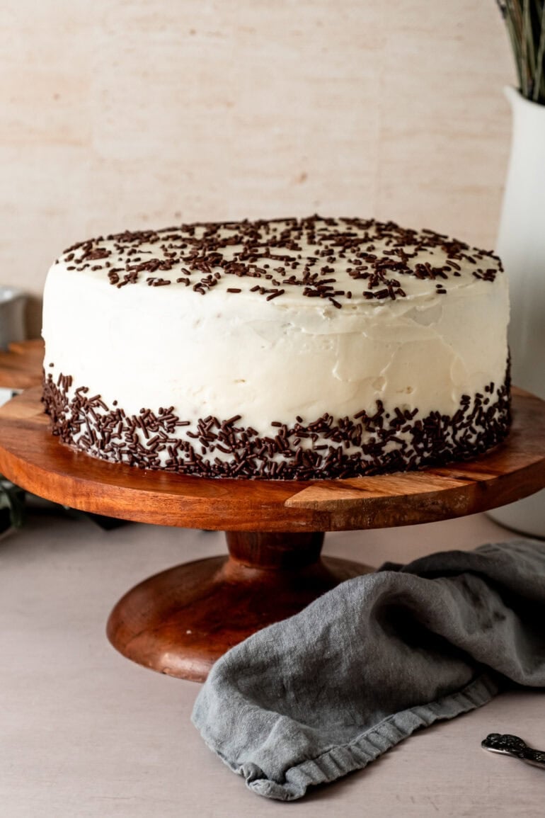 a two later chocolate cake covered with vanilla buttercream frosting and chocolate sprinkles on a wooden cake stand
