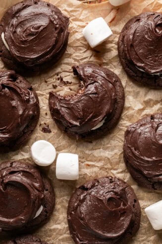 a photo of several chocolate marshmallow cookies with large marshmallows scatted around them sitting on a piece of brown parchment paper