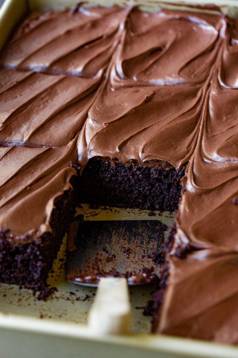 A pan of chocolate zucchini cake that has been cut into pieces and one piece has been removed. It is frosted with smooth, creamy chocolate frosting.