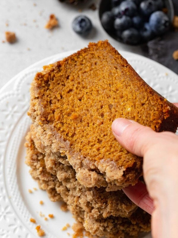 a photo of someone picking up a slice of pumpkin bread with a thick streusel topping from a pile of slices of bread.
