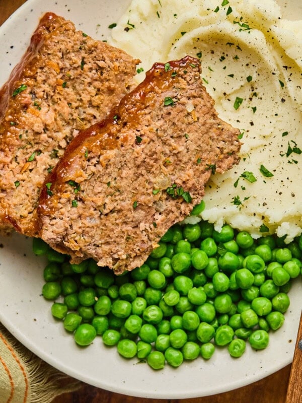 a photo of a plate full of two slices of meatloaf, some mashed potatoes and a serving of peas