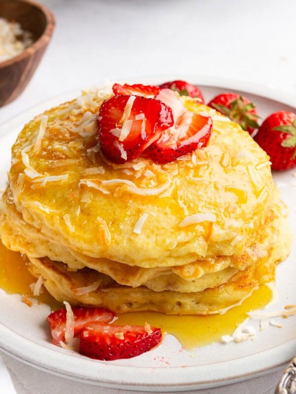 a stack of golden pancakes dripping with syrup and topped with toasted coconut and sliced strawberries