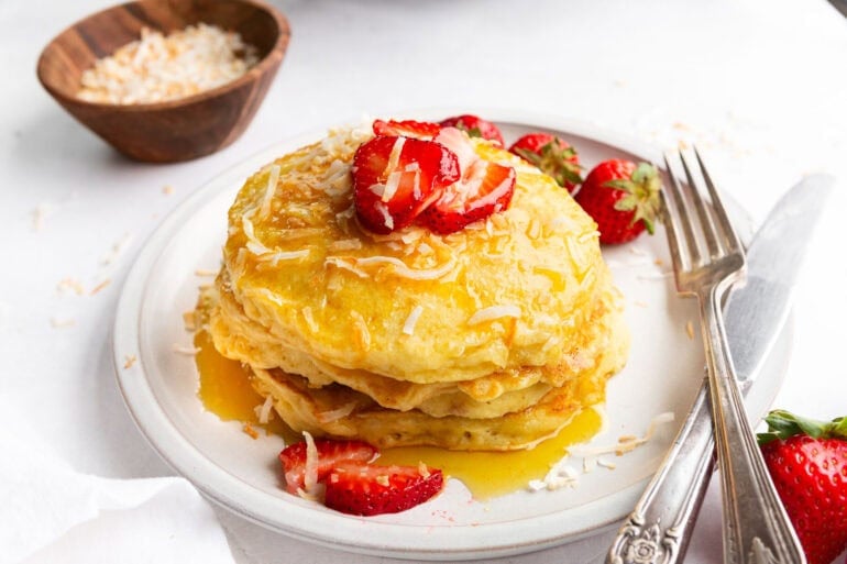 a stack of golden pancakes dripping with syrup and topped with toasted coconut and sliced strawberries
