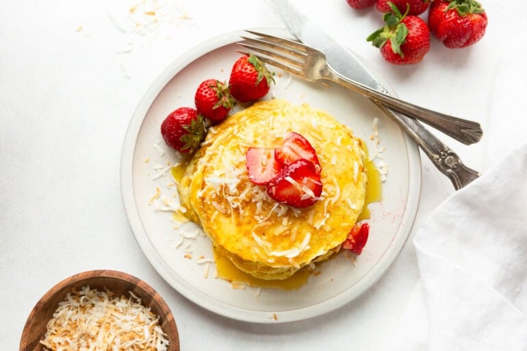 a plate with a stack of golden pancakes topped with toasted coconut, strawberry slices and for fork sitting next to the pancakes