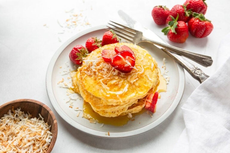 golden coconut pancakes stacked on a white plate topped with syrup, toasted coconut and sliced strawberries