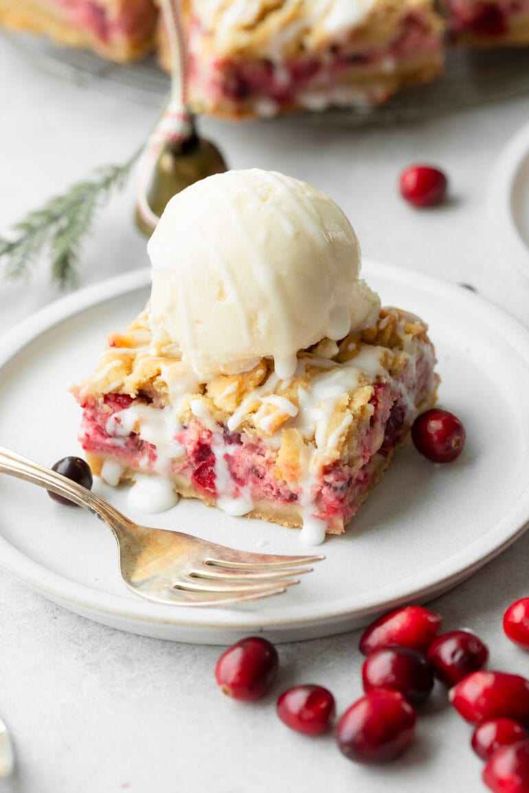 a photo of a cranberry crumb bar topped with a scoop of vanilla ice cream sitting on a white dessert plate with a fork sitting next to it