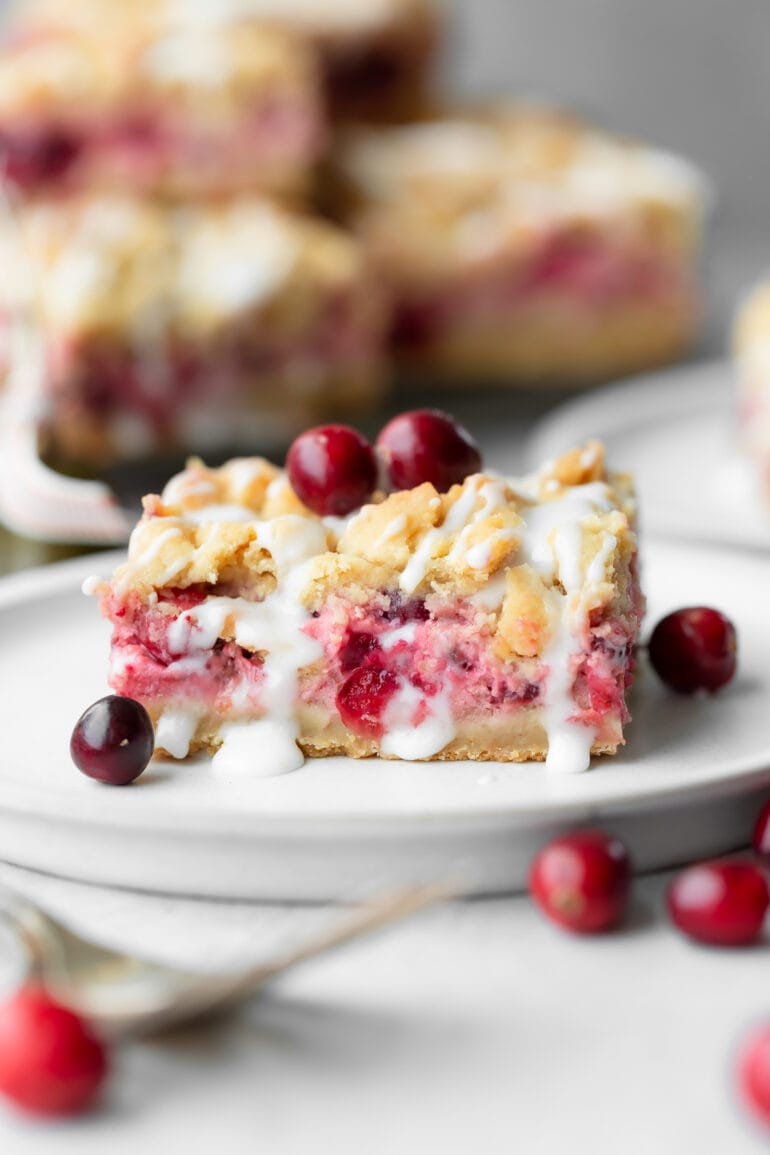 a photo of a cranberry crumb bar sitting on a small dessert plate drizzled with a white glaze and whole cranberries scattered around it