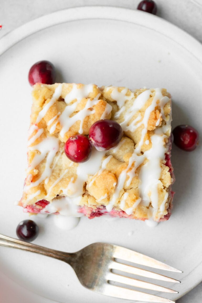a photo taken over the top of a single square cranberry crumb bar topped with a white glaze drizzle with two whole cranberries on top