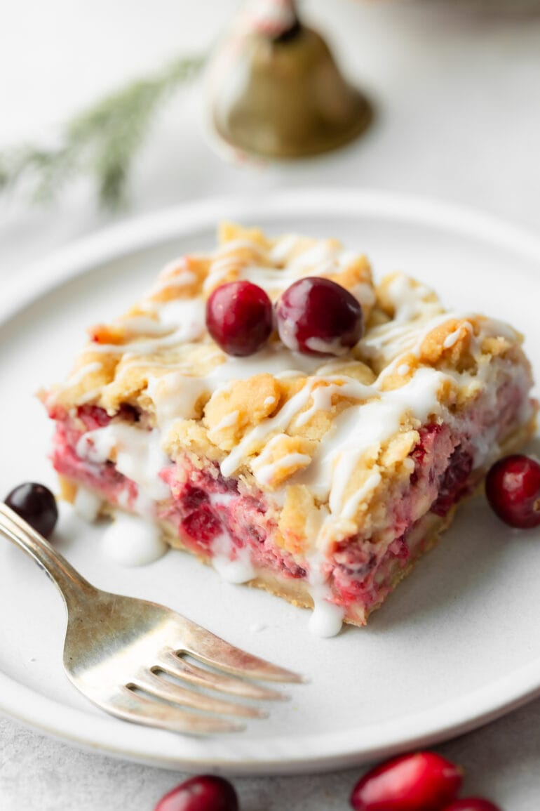 a photo of a cranberry bar sitting on a white dessert plate topped with a golden crumble and a white glaze