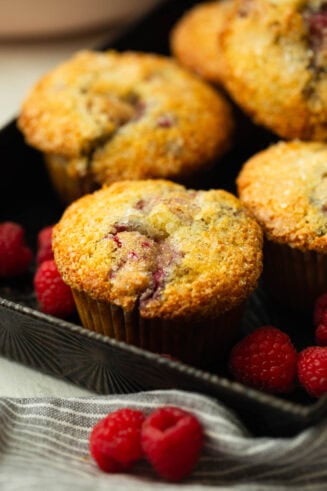 a photo of several golden baked raspberry muffins sitting in a serving tray surrounded by fresh raspberries