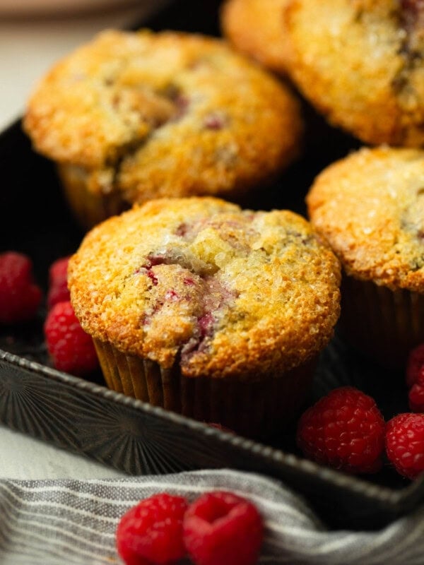 a photo of several golden baked raspberry muffins sitting in a serving tray surrounded by fresh raspberries