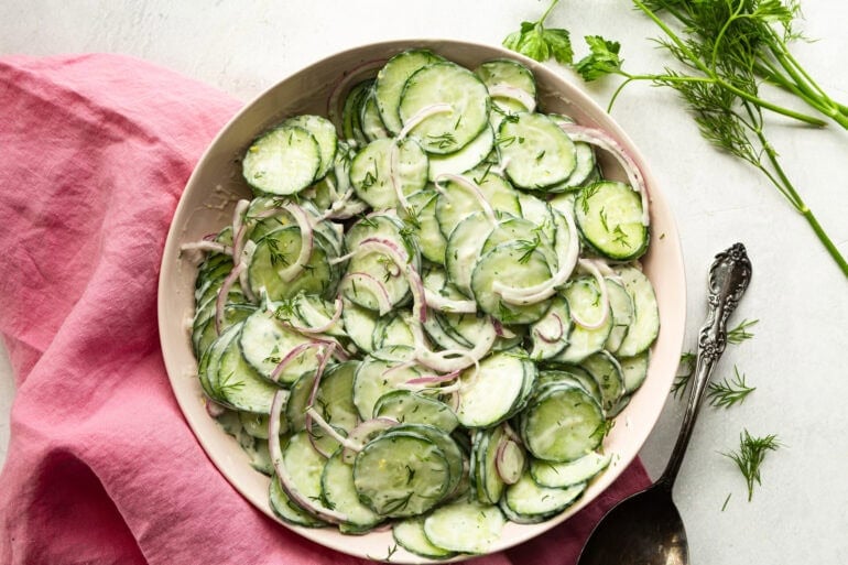 A close up of creamy cucumber salad showing thin cucumber slices and red onion tossed in a thick creamy dill dressing garnished with fresh basil.