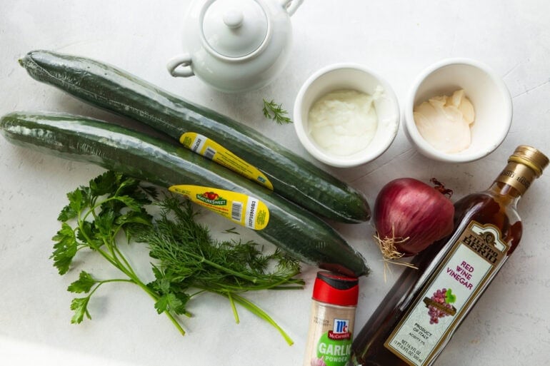 An overhead flat lay of creamy cucumber salad ingredients including whole English cucumbers, a bunch of fresh dill, a sliced red onion, and small bowls of sour cream and mayonnaise on a white marble surface.