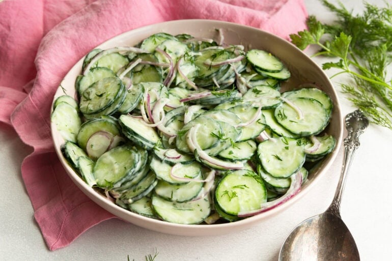 A side angle shot of a white serving bowl filled with veggies covered in a sour cream dressing sitting on a wooden surface with fresh dill and a linen napkin alongside.