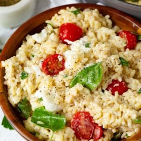 A bowl of caprese orzo salad. The salad has tomatoes, mozzarella chunks and fresh basil leave in it. A container of fresh ground pepper and a few parsley leaves are next to the salad.