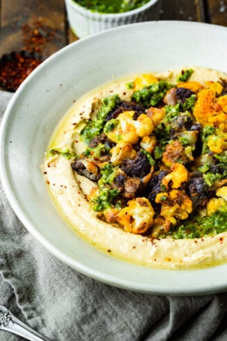 a phot of a serving bowl full of hummus topped with roasted tricolor roasted cauliflower and drizzled with pesto.