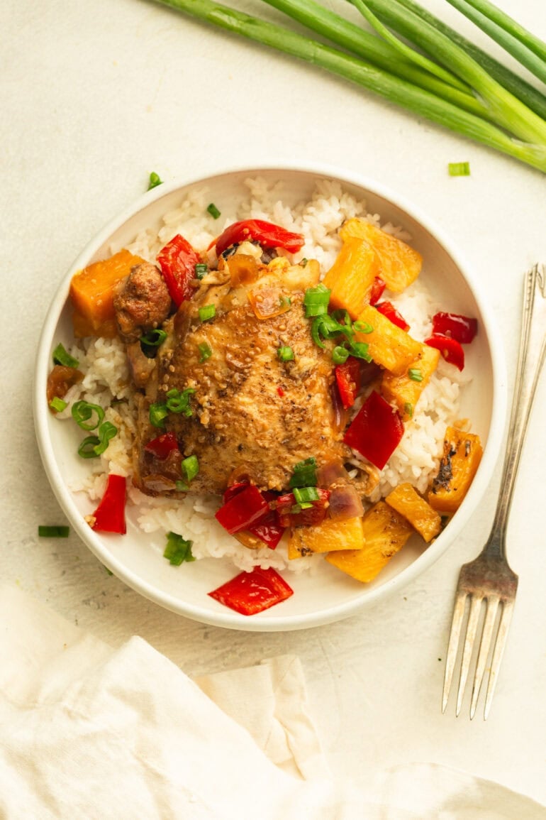 a white plate with a serving of white rice with a golden chicken thigh on top with saucy pineapple, red bell pepper and onions topped with chopped green onions