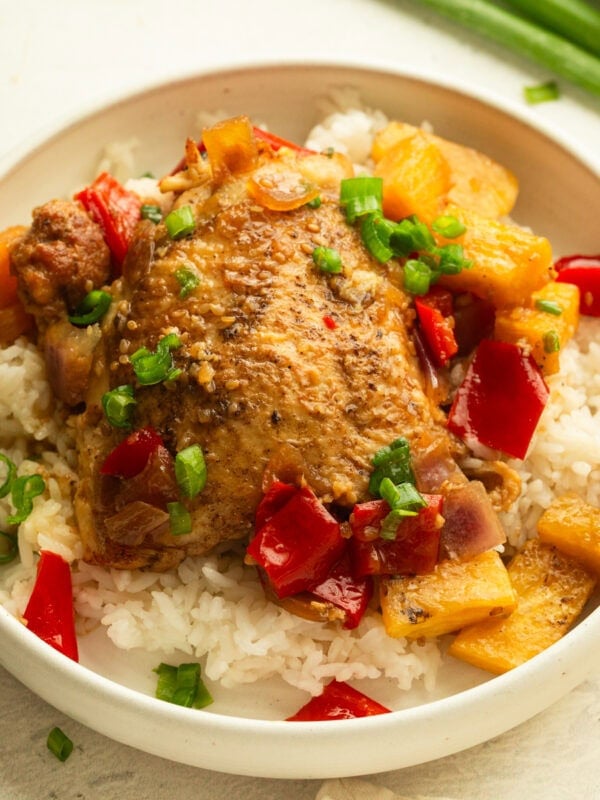 a serving of white rice topped with a golden chicken thigh, pineapple chunks, chopped red bell pepper and onions