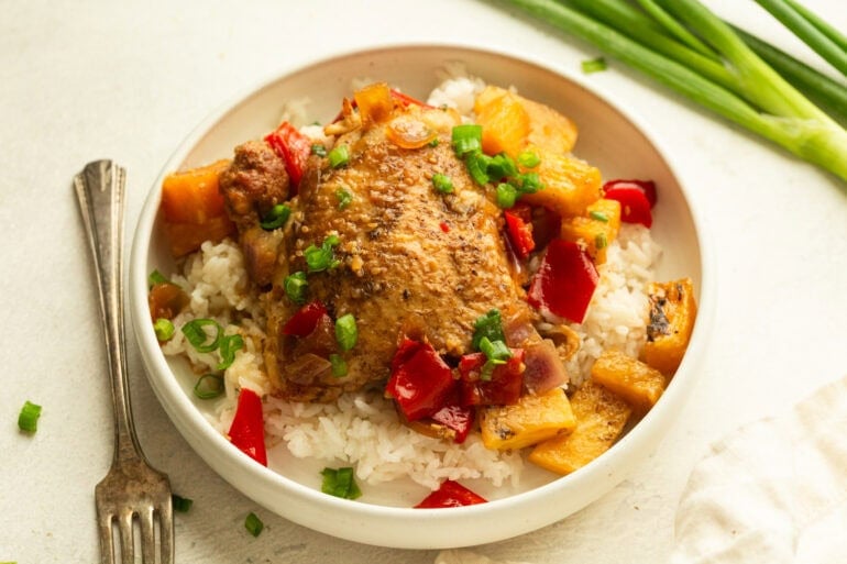 a serving of white rice topped with a golden chicken thigh, pineapple chunks, chopped red bell pepper and onions