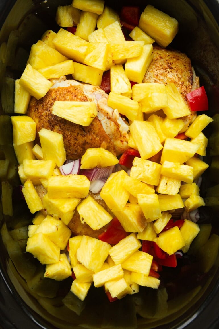 a black crockpot filled with pan seared chicken thighs, pineapple chunks, chopped red bell peppers and purple onion